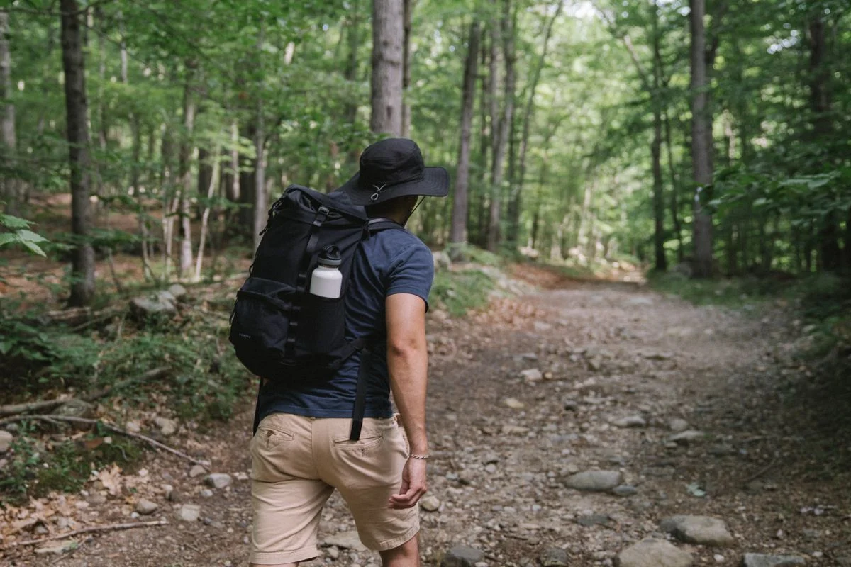 A man with a backpack hikes on a rocky forest path, wearing a hat.