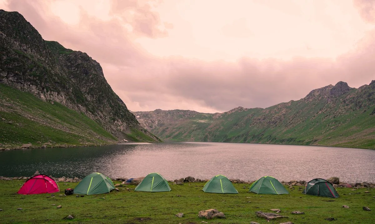 A serene campsite by a lake with tents on a grassy area, surrounded by mountains at dusk.