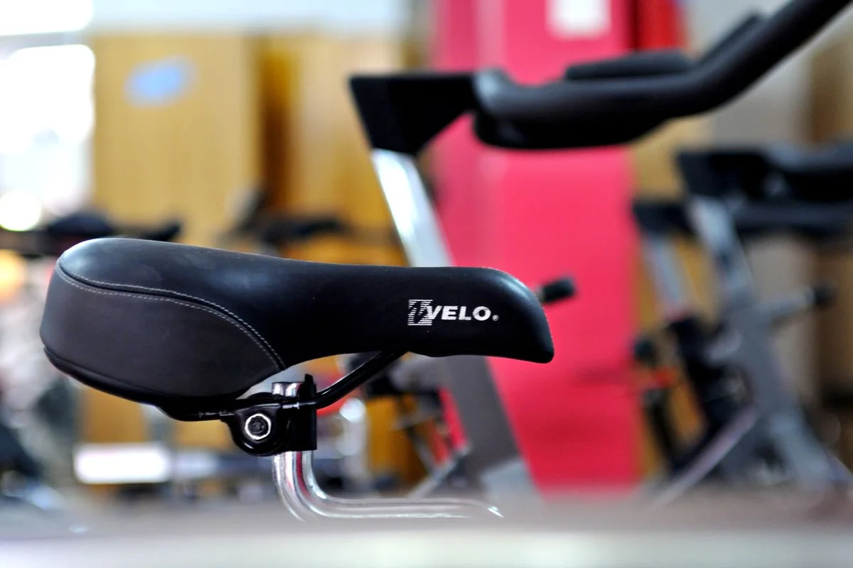 Detailed focus on a black bicycle saddle with gym equipment in the background.
