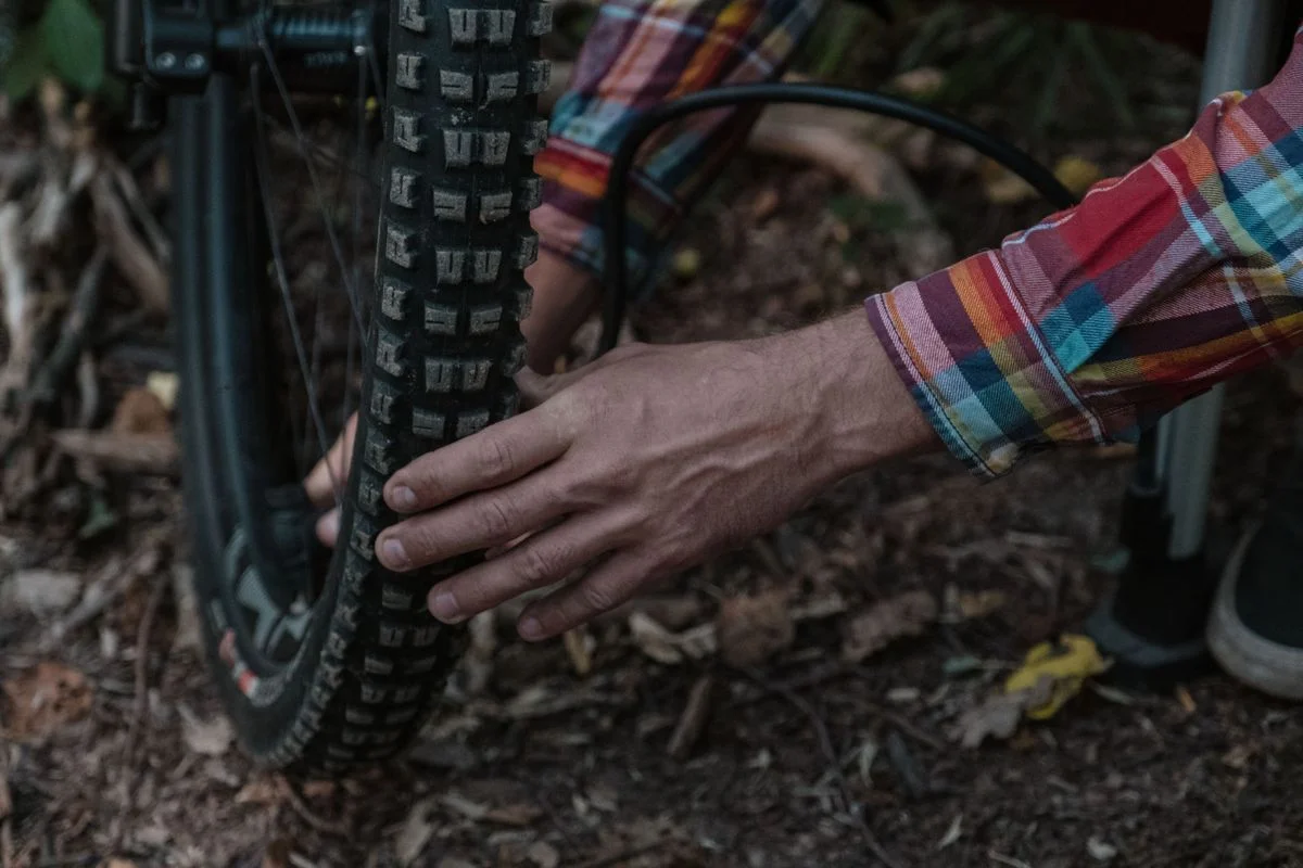 Person in plaid shirt inflating a mountain bike tire in a natural setting, highlighting maintenance and adventure.