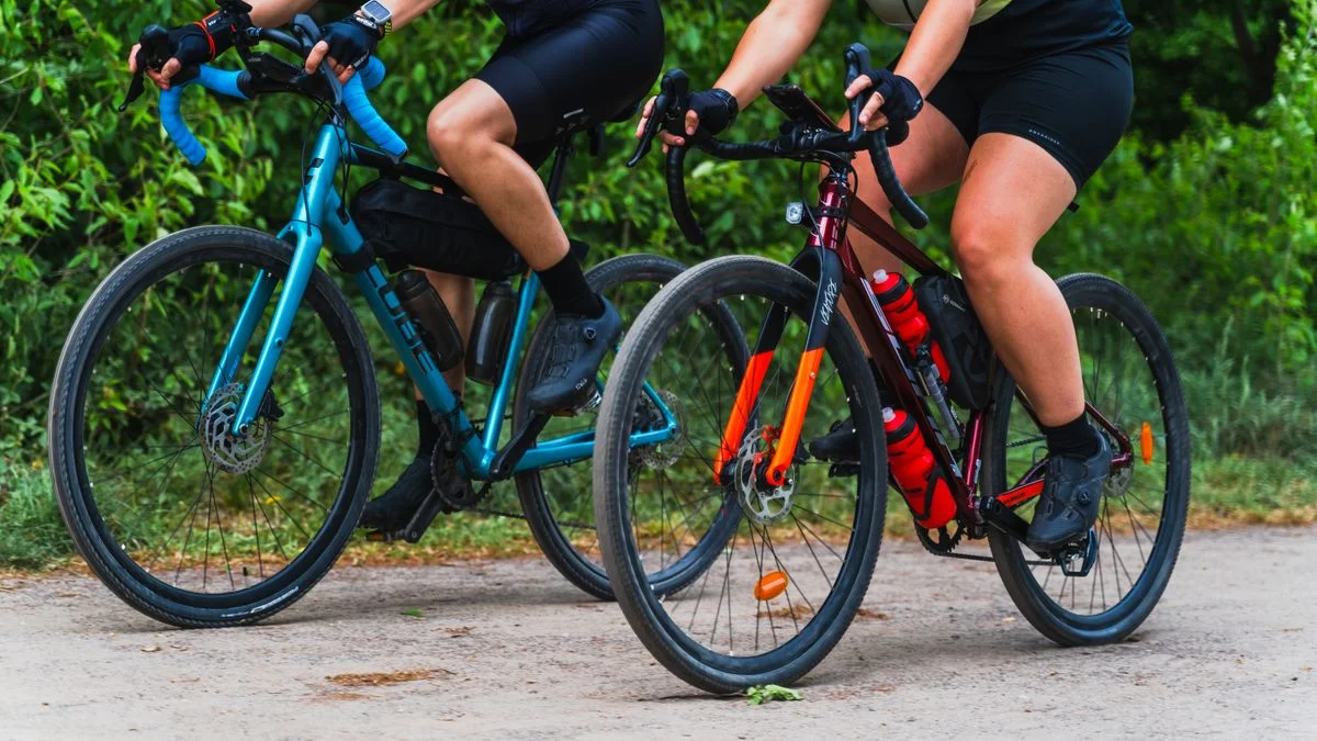 Two cyclists riding gravel bikes on a dirt path surrounded by greenery.